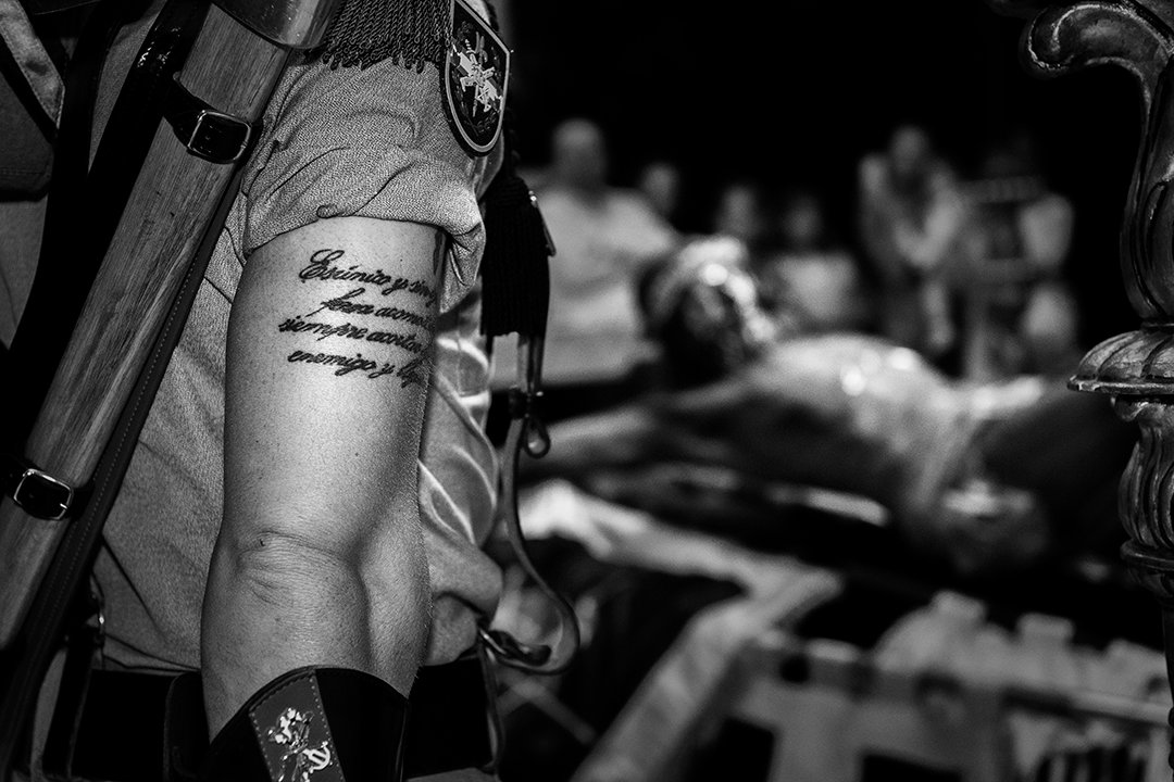 Detail of a tattooed arm with service members in the background.